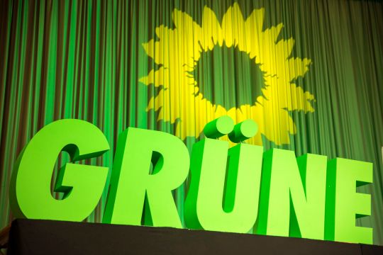 "GRÜNE" steht in großen Buchstaben auf einer Bühne, dahinter ist eine gelbe Sonnenblume auf den grünen Vorhang projiziert worden.