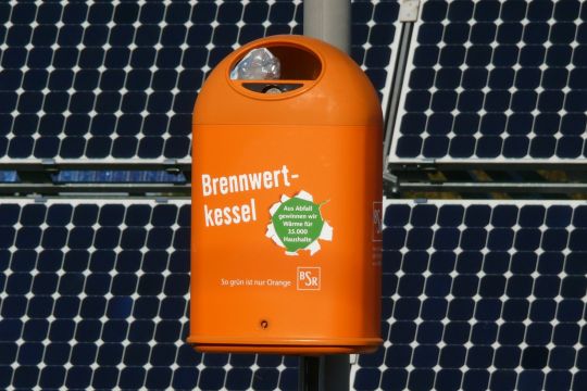 Öffentlicher Mülleimer der Berliner Stadtreinigung BSR mit der satirisch gemeinten Aufschrift: Brennwertkessel. Aus Abfall gewinnen wir Wärme für 35.000 Haushalte. Im Hintergrund Solarpaneele.