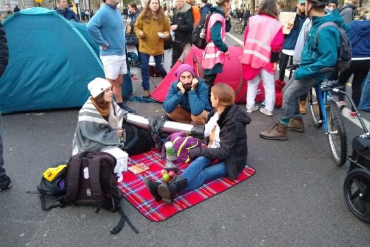 Zwei junge Frauen sitzen auf einer Decke auf der Straße und haben ihre Arme mit einem Lock-on verbunden.