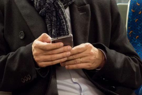 Mann im Anzug sitzt in der Bahn und schaut auf sein Smartphone, man sieht nur Hände und Oberkörper.