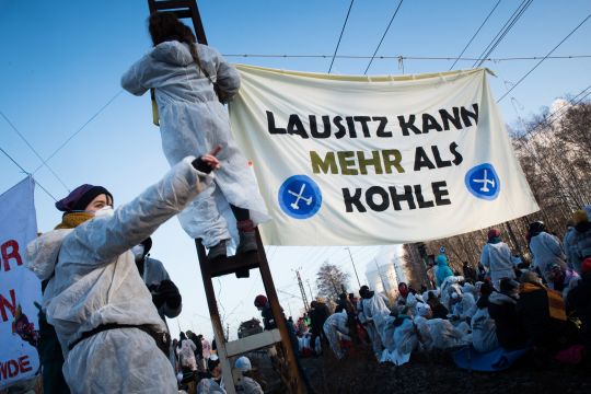 Weiß gekleidete Aktivisten stehen auf einem Bahngleis und hängen ein Transparent auf: 