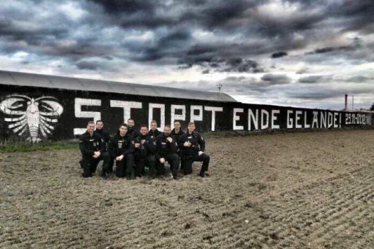 Eine Gruppe Uniformierter posiert vor einer schwarz gestrichenen Mauer mit der Aufschrift: 