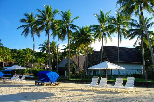 Leerer Strand mit einigen Liegestühlen und Sonnenschirmen, dahinter Palmen und Ferienhäuser, im Hintergrund eine bewaldete Anhöhe.