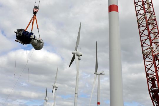Ein großer Stromgenerator hängt an einem Kranseil, daneben Kran und Windturm, im Hintergrund Windräder.
