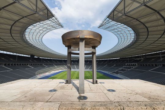 Steinschale auf Podest, im Hintergrund oval angeordneten Ränge und offenes Dach