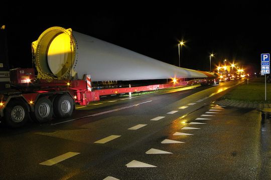 Auf einem überlangen Tieflader wird ein Windrotorblatt auf einer Straße durch die Nacht transportiert.