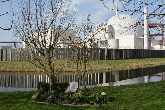 AKW-Anlage mit typischem halbkugelförmigem Reaktorgebäude, vor dem Sicherheitszaun an einem Baum ein mit Blumen umpflanzter Gedenkstein: 