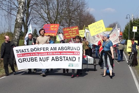 Kleine Demonstration gegen Braunkohle auf einer Dorfstraße mit bunten handgemalten Schildern und dem Transparent "Statt Dörfer am Abgrund – Grube auf Abstand!"