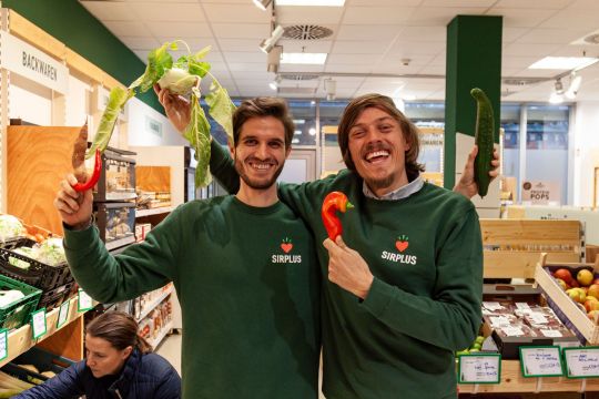 Martin Schott und Raphael Fellmer halten Gemüse hoch und lachen.