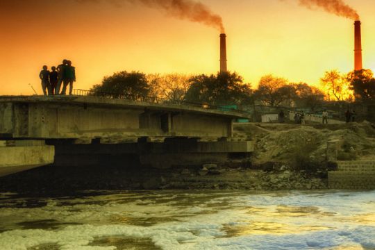 Verschmutzter Fluss mit Schaumkronen, im Hintergrund rauchende Fabrikschornsteine.