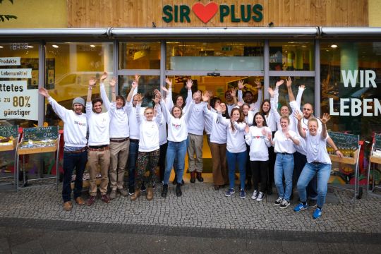 Das Team des Sirplus-Marktes in Berlin-Steglitz steht vor dem Laden und reckt die Arme in die Höhe.