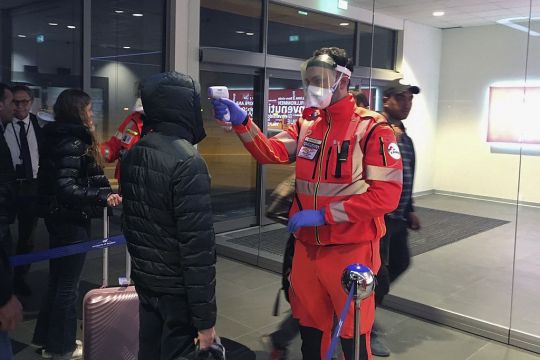 Ein Mann im Schutzanzug richtet ein pistolenartiges Prüfgerät auf die Stirn eines vor ihm stehenden Fluggastes.