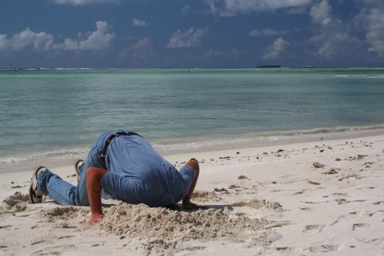 Mann kniet an Strand und hat seinen Kopf in den weißen Sand gesteckt