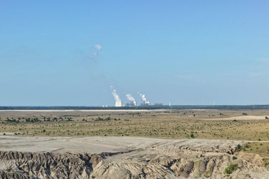 Riesige, spärlich bewachsene Tagebaufläche nach beendeter Förderung, am Horizont dampfende Kühltürme des Kohlekraftwerks.