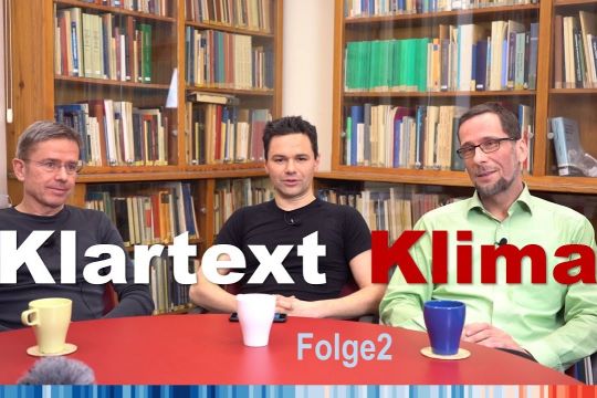 Özden Terli, Stefan Rahmstorf und Volker Quaschning unterhalten sich an einem Tisch, im Hintergrund Bücher, dazu der Schriftzug "Klartext Klima - Folge 2".