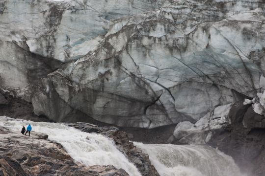 Zwei Menschen stehen am Rande eines reißenden Flüsschens in einer Stein- und Eislandschaft mit hoch aufragenden Wänden.