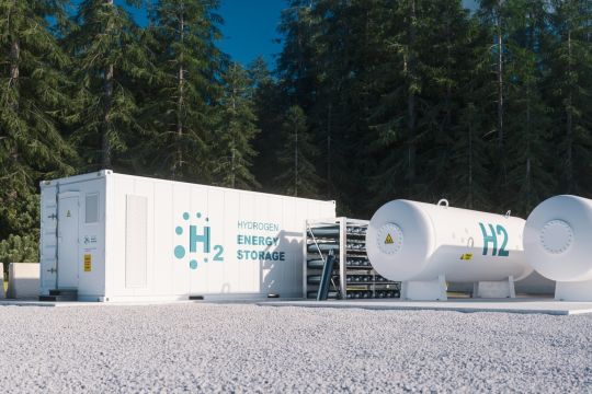 Weiße Container und Tanks mit der Aufschrift "H₂ Hydrogen Energy Storage", dahinter Wald.