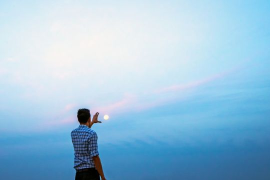 Mann steht vor dem Abendhimmel und greift den Mond scheinbar mit der Hand.