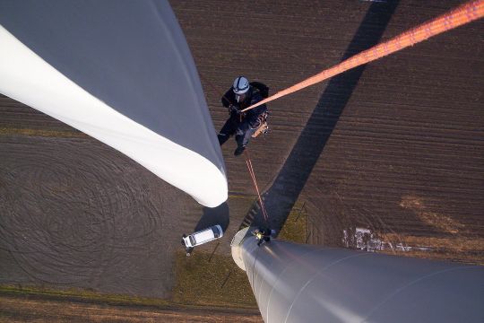 Aufnahme von oben an Windturm und Rotorblatt entlang nach unten, ein Monteur hängt am Seil, ganz klein ist am Boden ein Auto zu sehen.