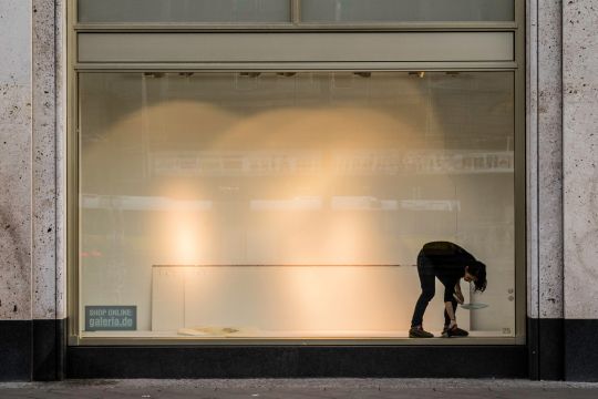 Leeres Schaufenster, in dem eine Angestellte aufräumt, auf einem kleinen Schild steht: "Shop online: galeria.de".