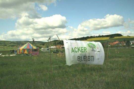 Auf einem Feld stehen ein großes buntes Zelt, ein Wohnwagen, Tripods und ein Transparent: "Der Acker bleibt!"