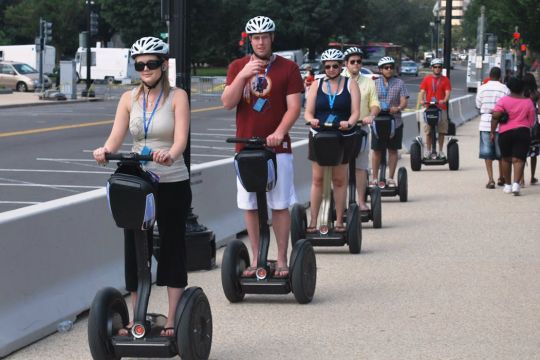 Touristische Gruppe rollt auf Segways hintereinander den Gehweg entlang, alle mit identischen Helmen und mit Badges um den Hals.