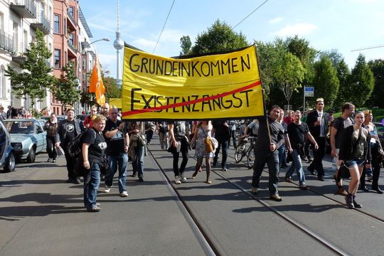 Viele Menschen auf Demo bei gutem Wetter, Fronttransparent mit Aufschrift "Grundeinkommen" und einem durchgestrichenen "Existenzangst", Am Rand Altbau-Häuserzeile