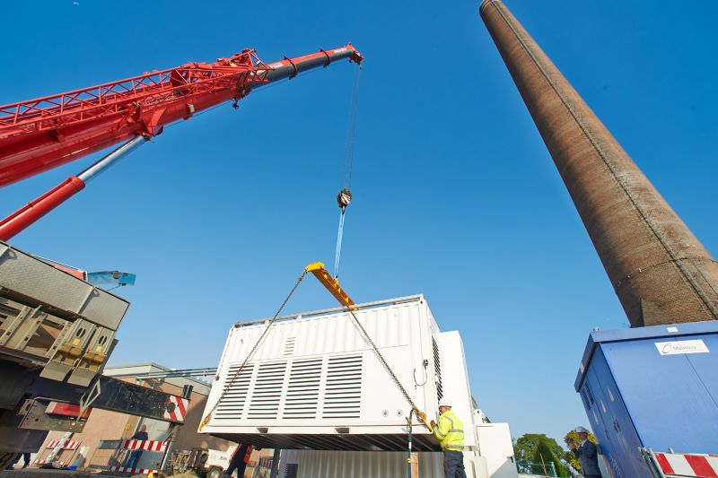 Kran hebt Container mit Wasserstoff-Elektrolyseur auf Platz neben einem alten Beton-Schornstein.