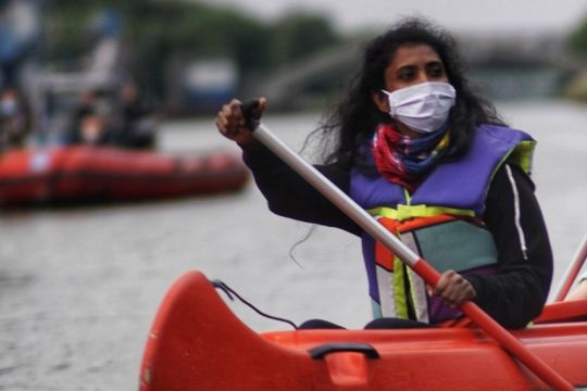 Frau mit Mund-Nasen-Schutz in Boot, im Hintergrund verschwommen noch ein Boot und das Kraftwerk Datteln