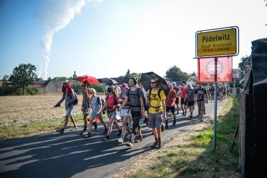 Eine Gruppe von Klimaaktivisten geht auf einer Straße, zu sehen ist das Ortsschild von Pödelwitz und im Hintergrund ein Kraftwerk. 