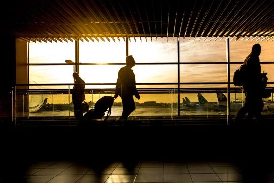 Reisende in der Abfertigungshalle eines Flughafens, durch das große Fenster ist zu sehen, wie die tief stehende Sonne auf das Rollfeld scheint.