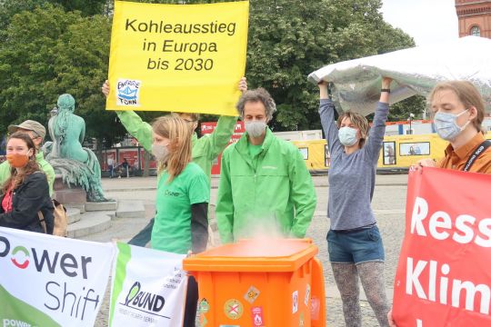 Aus einer orangefarbenen Mülltonne steigt Rauch auf, dahinter demonstrieren Jugendliche mit Schildern für Klimaschutz.