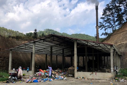 In einer seitlich offenen Halle wird Plastikmüll verbrannt, aus dem Schornstein kommt dunkler Rauch.