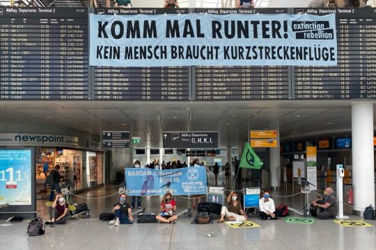 Einige Menschen sitzen vor der Anzeigetafel des Flughafens München, über der Tafel hängt ein Transparent: "Komm mal runter! Kein Mensch braucht Kurzstreckenflüge".