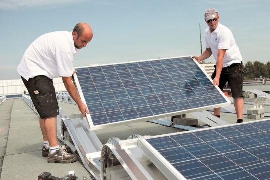 Zwei Monteure installieren ein Solarmodul auf dem Flachdach eines Hauses.
