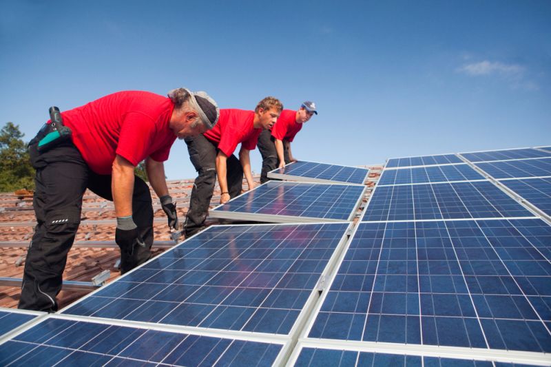 Drei Monteure in roten T-Shirts installieren Solarmodule auf einem Dach in Landshut (Niederbayern).