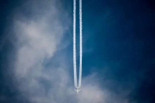 Über einen teils wolkigen, teils verdunkelten Himmel fliegt ein Flugzeug von oben nach unten mit einem weißen Kondensstreifen durchs Bild.