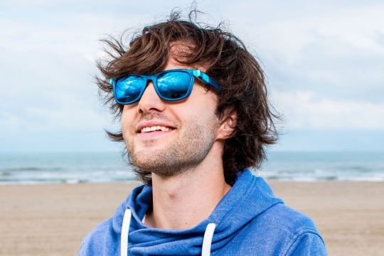 Boyan Slat steht am Strand und lächelt hinter einer blau verspiegelten Brille und schaut dabei leicht nach oben.