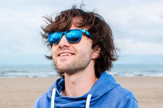 Boyan Slat steht am Strand und lächelt hinter einer blau verspiegelten Brille und schaut dabei leicht nach oben.