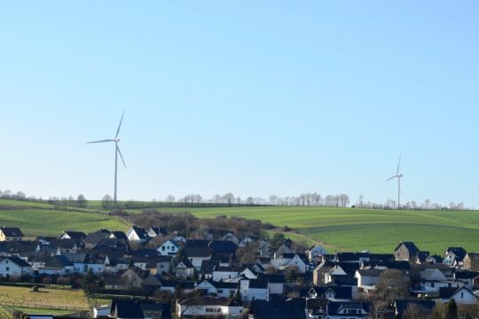 Ansicht von Ferne auf fast ein ganzes Dorf, hinten am Horizont der welligen Landschaft stehen zwei Windräder.