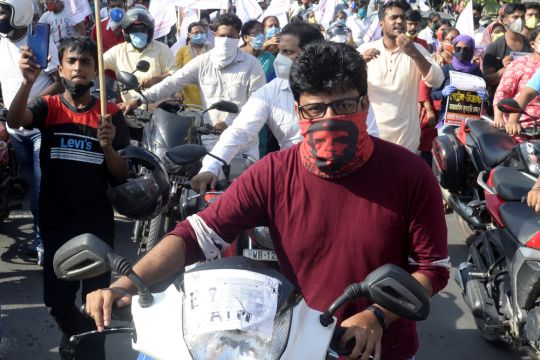 Männer schieben ihre Motorräder auf einer Demonstration in Kalkutta, der vordere schaut ernst, er trägt als Mundschutz ein Tuch mit Che-Guevara-Porträt.