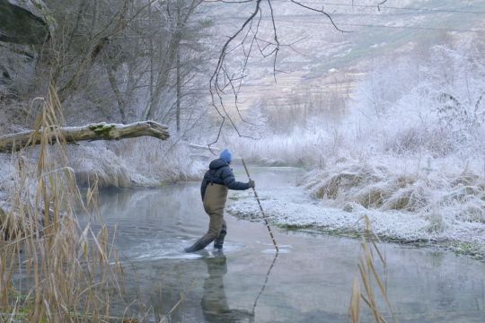 Ein Mensch in Wathose läuft mit einer Stocherstange durch die winterliche Rhône, die hier noch schmal und flach ist.