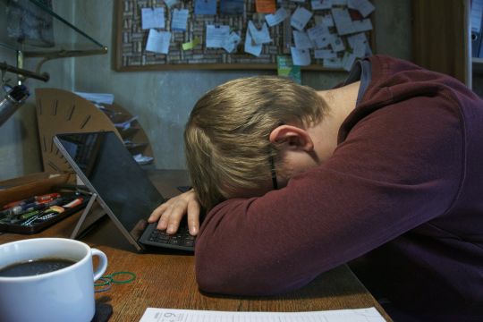 Junger Mann ist vor seinem aufgeklappten Laptop eingeschlafen, neben ihm eine große Tasse mit Kaffee.