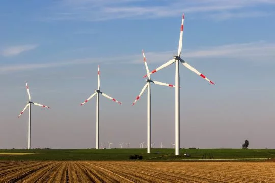Vier mittelgroße Windkraftanlagen auf einem Acker in ebener Landschaft, ganz im Hintergrund ein weiterer Windpark.
