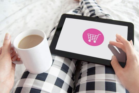Person in karierter Schlafanzughose mit Kaffeetasse und Tablet, auf dem ein Einkaufswagen-Icon zu sehen ist.