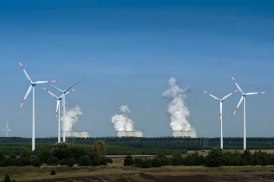 Einige Windräder in Landschaft mit Wäldern und feldern, im Hintergrund ein großes Kohlekraftwerk mit neun dampfenden Kühltürmen.