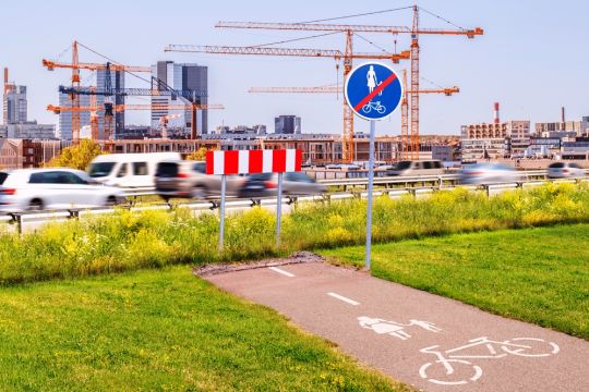 Ein Fuß- und Radweg endet plötzlich an einer stark befahrenen Schnellstraße, dahinter bauen Kräne eine moderne Stadt mit Hochhäusern.