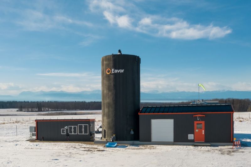 Ein schwarzer Silo-artiger Turm mit der Aufschrift Eavor, daneben ein Garagen-artiges, schwarzes Gebäude und ein schwarzer Container, alles in einer winterlichen Ebene.