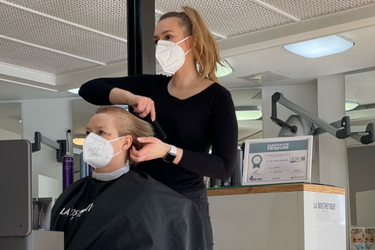 Friseurin frisiert Kundin, beide mit medizinischer Maske, im Hintergrund ein Schild: Klimapositive Friseure.