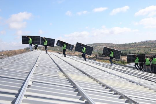 Monteure tragen an einem sonnigen Tag Solarpaneele auf das Dach eines größeren Gebäudes in Palästina. 